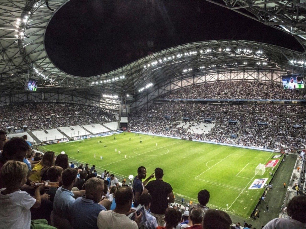 Fans in the stands watching a match under floodlights at a packed football stadium